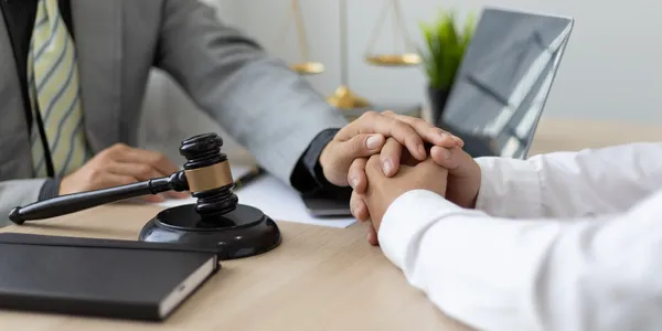 Two people sit at a desk in a law firm, one offering comfort by holding the other's hands. A judge's gavel and notebook are visible on the table.