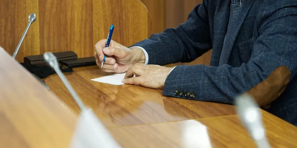 A person in a suit jacket is writing on a piece of paper at a wooden desk, possibly preparing documents for a law firm.