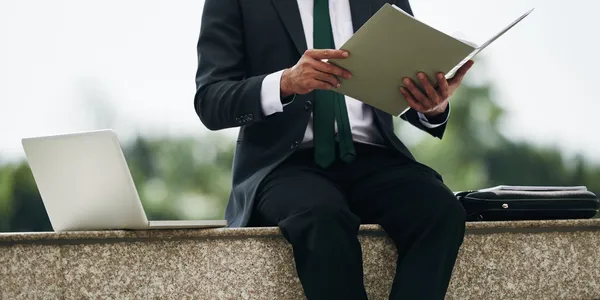 A person in a suit sits on a stone ledge outdoors, holding a tablet, with a laptop and briefcase beside them—likely preparing documents for their law firm—with city buildings in the background.