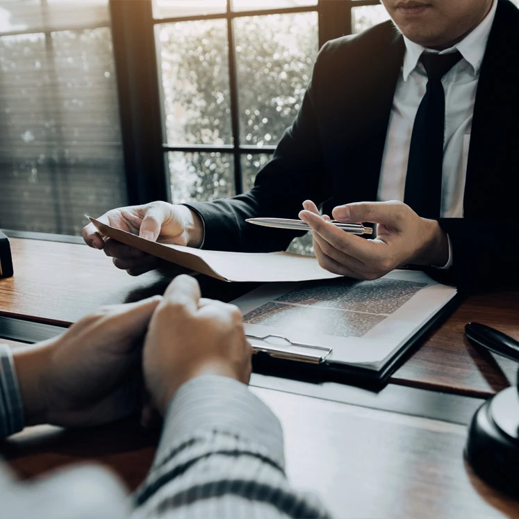 Two people in business attire sit at a desk with documents, one holding a pen and folder, appearing to discuss paperwork with a civil law attorney in a professional setting.